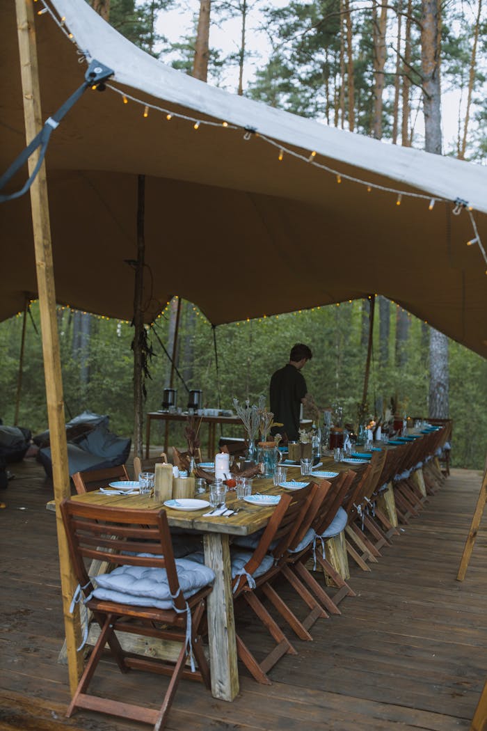 gallery-2 A beautifully arranged rustic outdoor dining setup under a tent in a wooded area, creating a cozy ambiance.